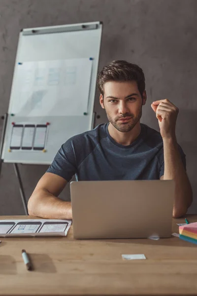 Enfoque selectivo del diseñador guapo mirando a la cámara por ordenador portátil y bocetos web móviles en la mesa - foto de stock