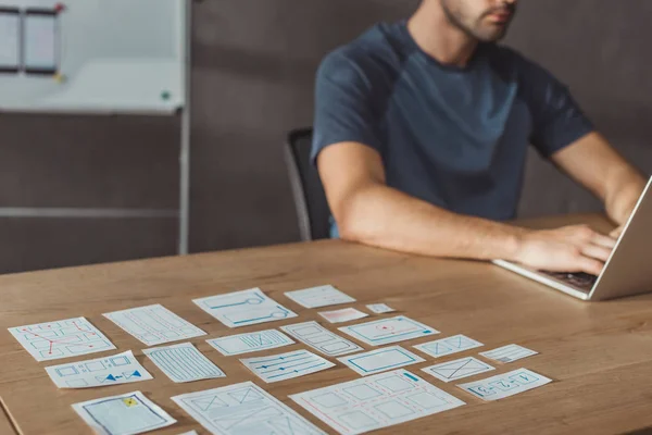 Vue recadrée du développeur ux à l'aide d'un ordinateur portable à côté des croquis du concepteur sur la table, mise au point sélective — Photo de stock