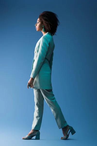 Vista laterale di attraente donna afro-americana in abito camminare su sfondo blu — Foto stock