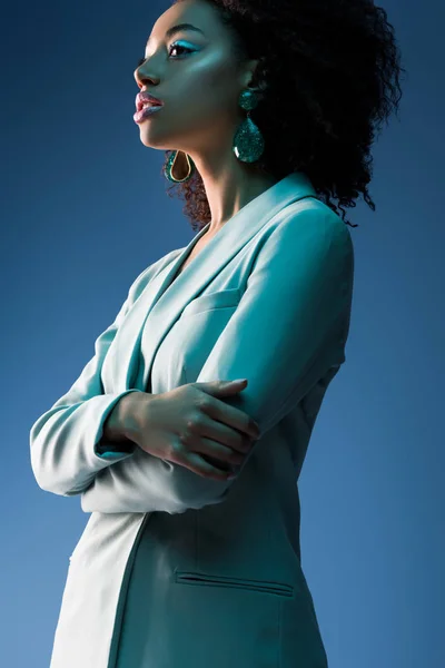 Atractiva mujer afroamericana en traje con brazos cruzados aislados en azul - foto de stock