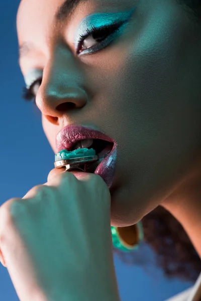 Atractiva mujer afroamericana con anillo mordedor de maquillaje aislado en azul - foto de stock