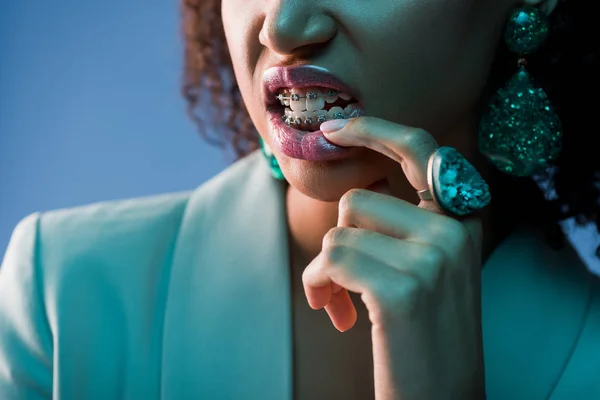 Обрезанный вид на африканскую американскую женщину с брекетами кусающий палец изолирован на синий — стоковое фото