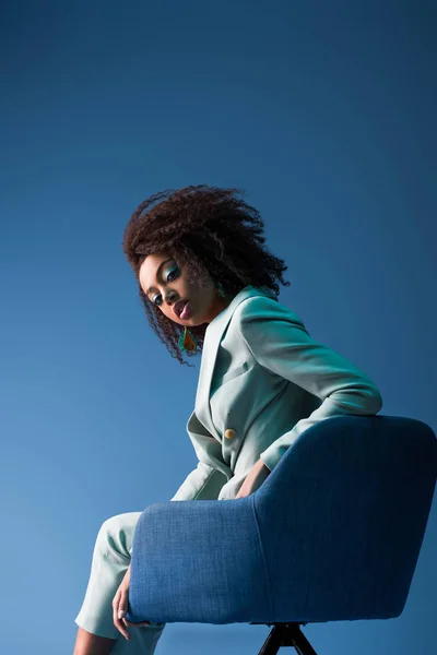 African american woman sitting on armchair isolated on blue — Stock Photo