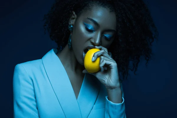 Atractiva mujer afroamericana con maquillaje comer limón aislado en azul - foto de stock