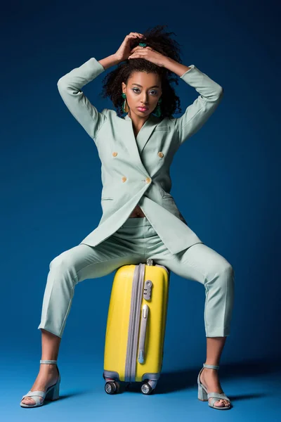 Atractiva mujer afroamericana sentada en una bolsa de viaje sobre fondo azul - foto de stock