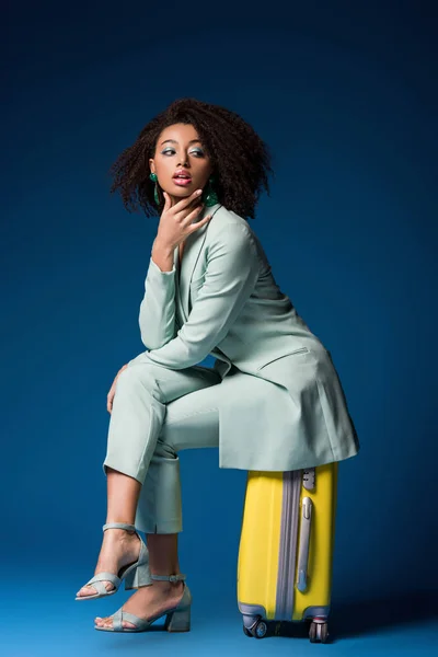 Atractiva mujer afroamericana sentada en una bolsa de viaje sobre fondo azul - foto de stock