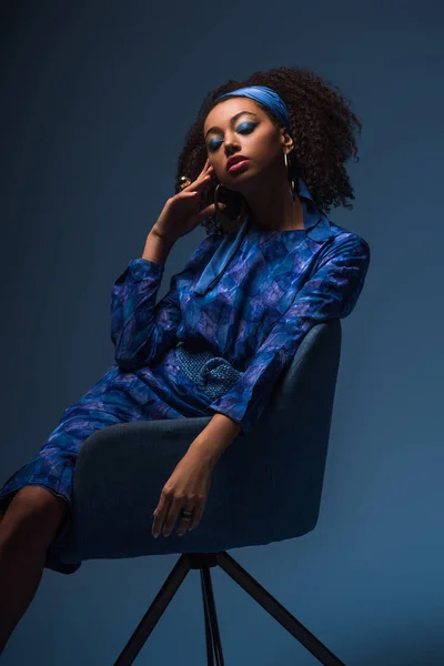 Attrayant afro-américain femme avec les yeux fermés assis sur un fauteuil isolé sur bleu — Photo de stock