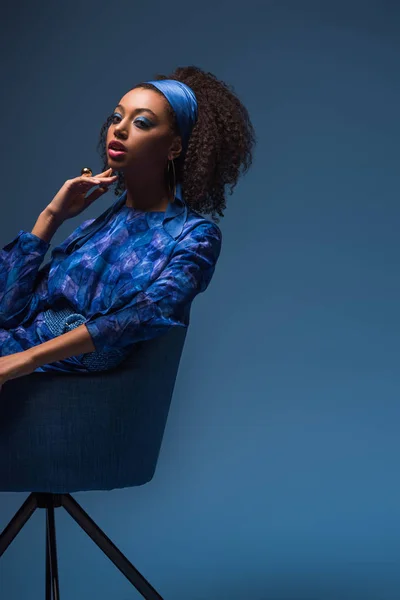 Attractive african american woman sitting on armchair isolated on blue — Stock Photo