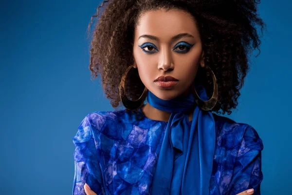 Stylische afrikanisch-amerikanische Frau in Kleid und Ohrringen, die isoliert auf blau in die Kamera schaut — Stockfoto