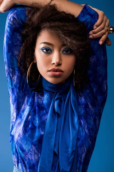 Mujer afroamericana con estilo con maquillaje en vestido mirando a la cámara aislada en azul - foto de stock