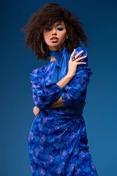Atractiva mujer afroamericana en vestido mirando a la cámara aislada en azul - foto de stock