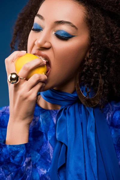 Mujer afroamericana con maquillaje comiendo sabroso limón aislado en azul - foto de stock