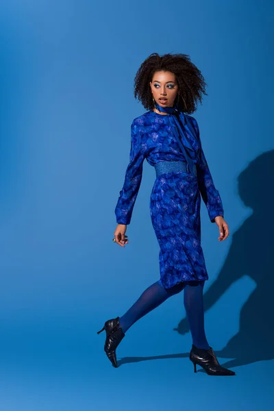 Séduisante femme afro-américaine en robe marchant sur fond bleu — Photo de stock