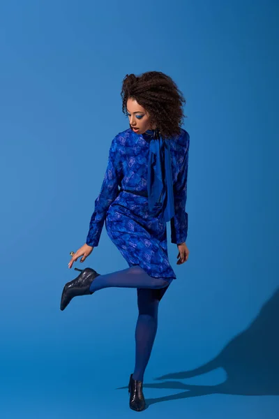 Attractive african american woman in dress touching her footwear on blue background — Stock Photo