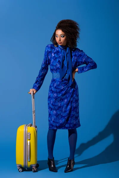 Afrikanisch-amerikanische Frau mit Hand an der Hüfte hält Reisetasche auf blauem Hintergrund — Stockfoto