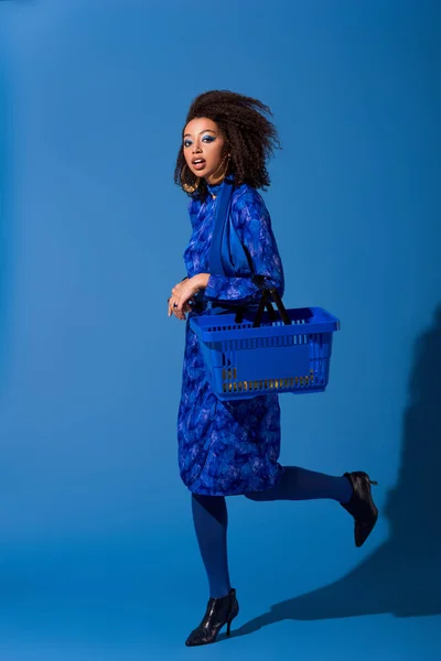 African american woman holding shopping basket on blue background — Stock Photo