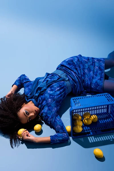 Mulher afro-americana deitada com cesta de compras e limões em fundo azul — Fotografia de Stock