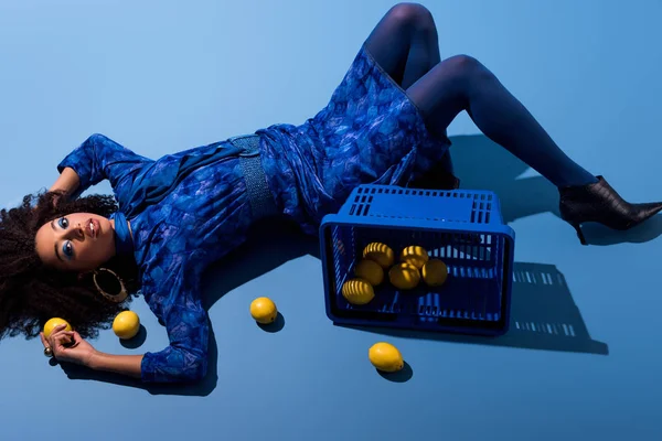 African american woman lying with shopping basket and lemons on blue background — Stock Photo