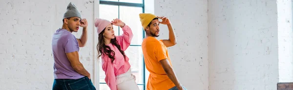 Panoramic shot of young multicultural dancers touching hats while breakdancing — Stock Photo