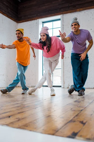 Menina elegante gesticulando enquanto breakdancing com homens multiculturais felizes em chapéus — Fotografia de Stock