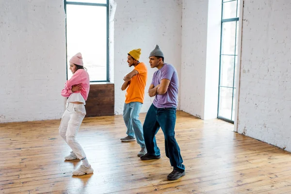 Stylish girl with crossed arms breakdancing with handsome multicultural men in hats — Stock Photo