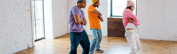 Panoramic shot of stylish girl with crossed arms breakdancing with handsome multicultural men in hats — Stock Photo