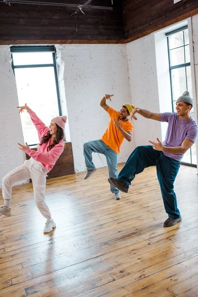 Attractive girl breakdancing and gesturing with stylish multicultural men in hats — Stock Photo