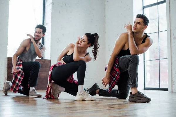 Hombres multiculturales guapos y mujer atractiva de pie de rodillas mientras bailan jazz funk - foto de stock