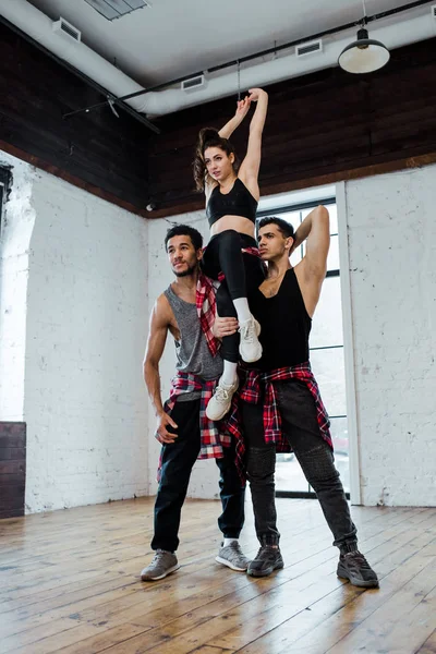 Strong multicultural men holding attractive woman while posing in dance studio — Stock Photo