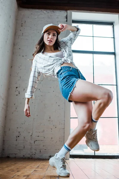 Attractive dancer posing and touching cap in dance studio — Stock Photo