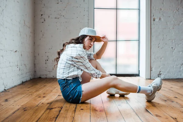 Beautiful dancer touching cap and sitting on floor in dance studio — Stock Photo