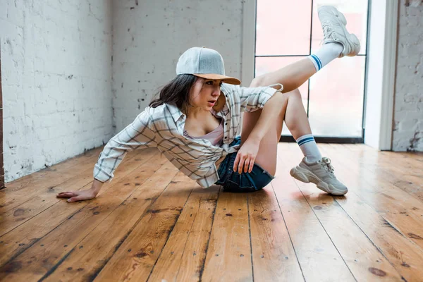 Beautiful dancer in cap sitting on floor and dancing hip-hop in dance studio — Stock Photo