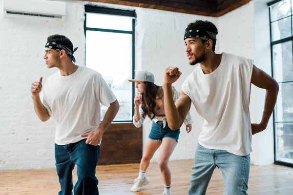 Мультикультурні чоловіки в головних уборах і приваблива жінка в кепці танцює хіп-хоп — стокове фото