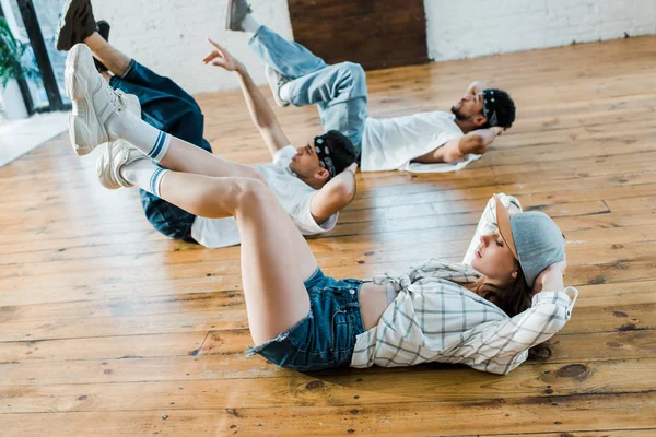Вибірковий фокус модної дівчини в кепці лежить на підлозі з мультикультурними танцюристами в танцювальній студії — Stock Photo