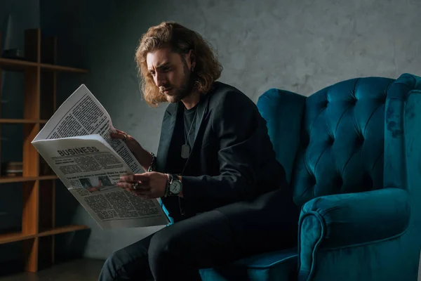 Напружений модний бізнесмен в чорному костюмі читає газету в кріслі в темряві — стокове фото