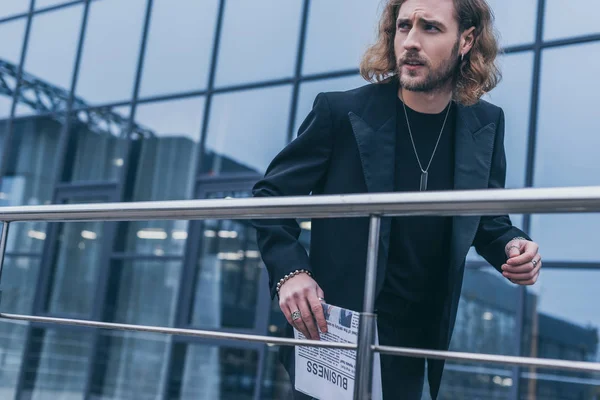 Hombre de negocios de moda en traje negro caminando con periódico cerca del edificio de oficinas y barandilla — Stock Photo