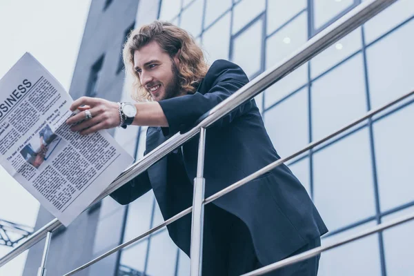 Vista de ángulo bajo de sonriente hombre de negocios de moda en traje negro leyendo periódico de negocios cerca de edificio de oficinas y barandilla - foto de stock