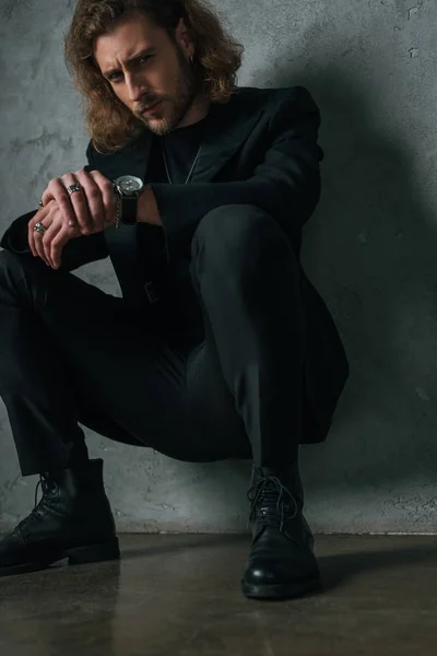 Напружений модний бізнесмен в чорному костюмі, сидячи в темряві біля сірої стіни — стокове фото