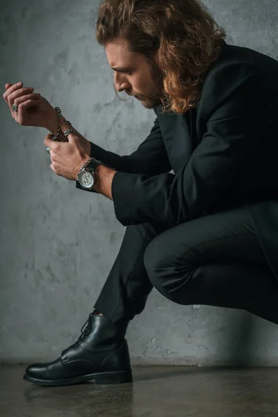 Модный бизнесмен в черном костюме трогает браслет в темноте возле серой стены — стоковое фото