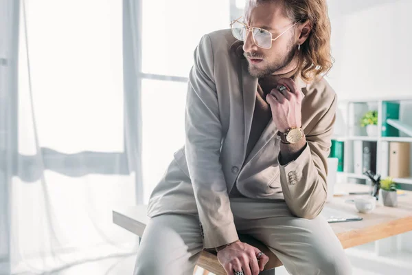 Low angle view of elegant fashionable businessman sitting on desk and looking away in office — Stock Photo