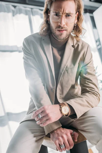 Low angle view of elegant fashionable businessman sitting on desk and looking at camera in office — Stock Photo