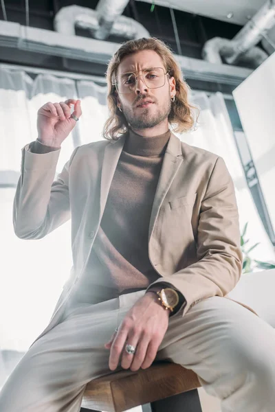 Low angle view of elegant fashionable businessman sitting on desk and looking at camera in office — Stock Photo