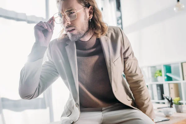 Low angle view of elegant fashionable businessman sitting on desk and looking away in office — Stock Photo