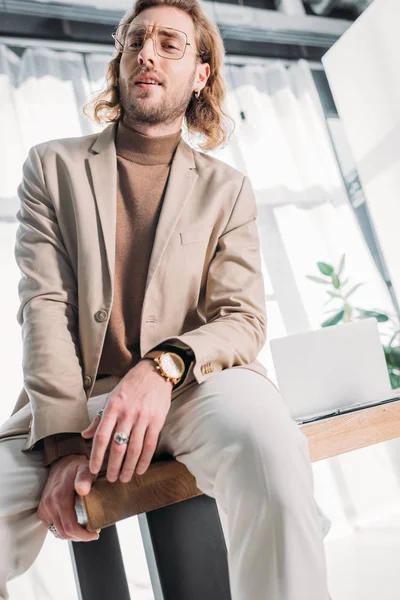 Низький кут зору впевнений модний бізнесмен сидить на столі і позує в офісі — стокове фото