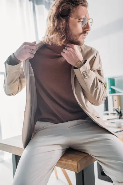 Vista angolo basso di elegante uomo d'affari alla moda seduto sulla scrivania e in posa in ufficio — Foto stock