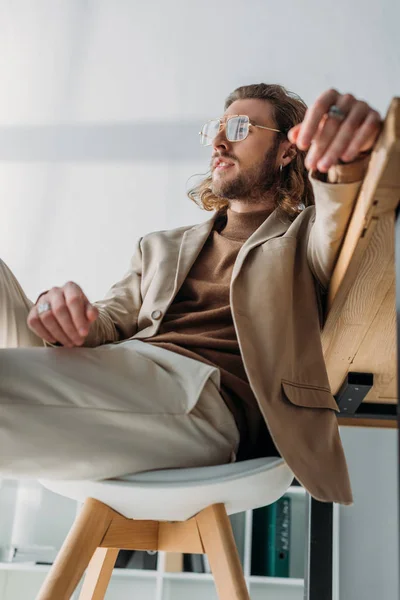 Elegante hombre de negocios de moda sentado en la silla cerca del escritorio en la oficina - foto de stock