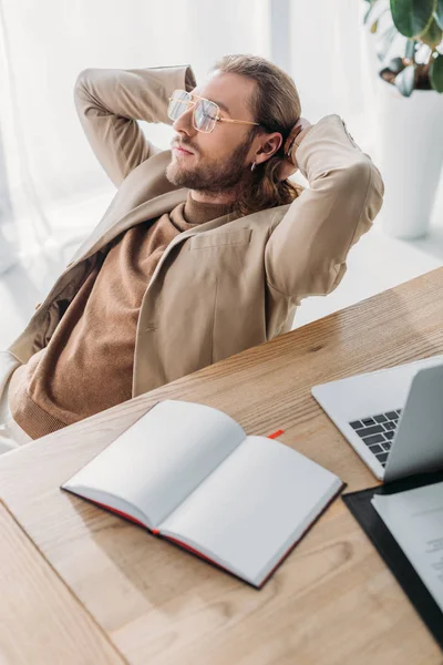 Selective focus of elegant fashionable businessman sitting on chair with closed eyes and hands behind head near desk with blank notebook in office — Stock Photo
