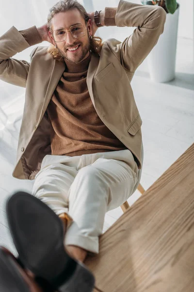 Enfoque selectivo de hombre de negocios de moda feliz sentado en la silla con las piernas en el escritorio y las manos detrás de la cabeza en la oficina - foto de stock