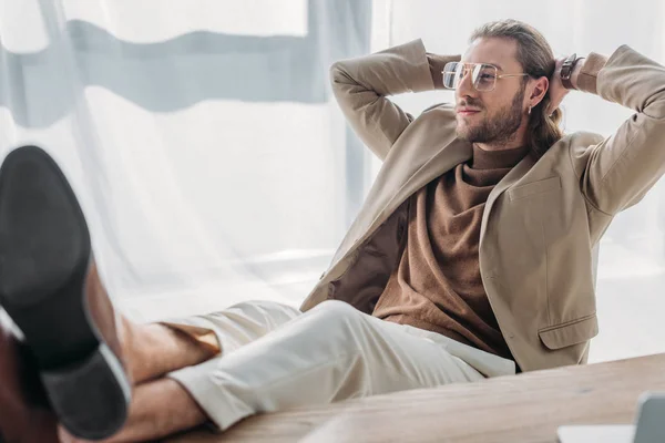 Selektiver Fokus eines eleganten modischen Geschäftsmannes, der auf einem Stuhl sitzt, mit Beinen auf dem Schreibtisch und Händen hinter dem Kopf im Büro — Stockfoto