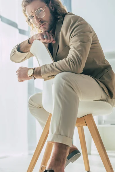 Elegante uomo d'affari alla moda in posa sulla sedia e guardando la fotocamera alla luce del sole in ufficio — Foto stock